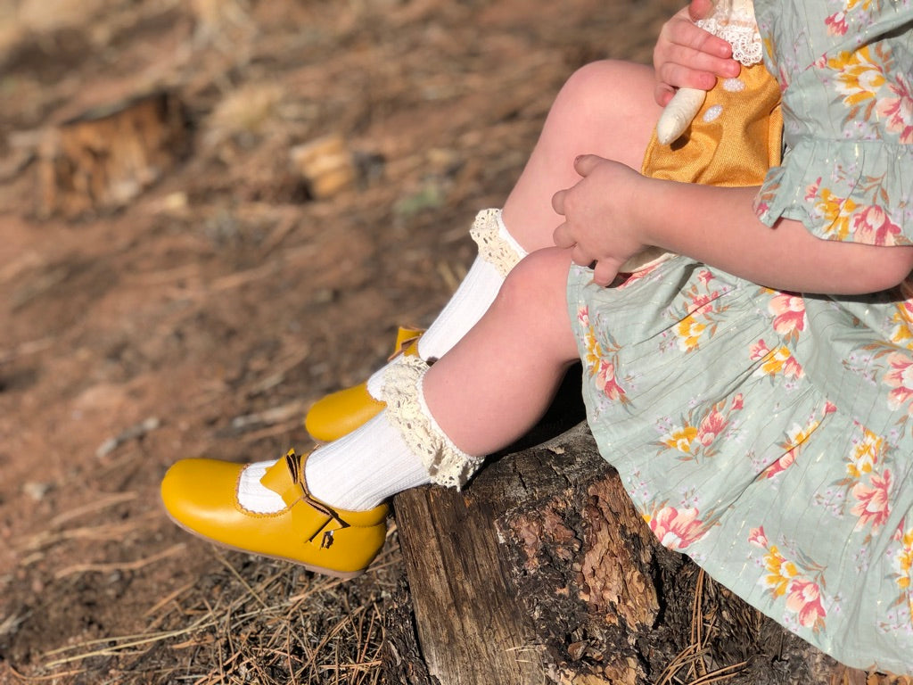 Toddler in floral dress and lace‑trim socks wearing mustard yellow Mary Jane shoes, sitting on a tree stump holding a small doll in a rustic outdoor setting.