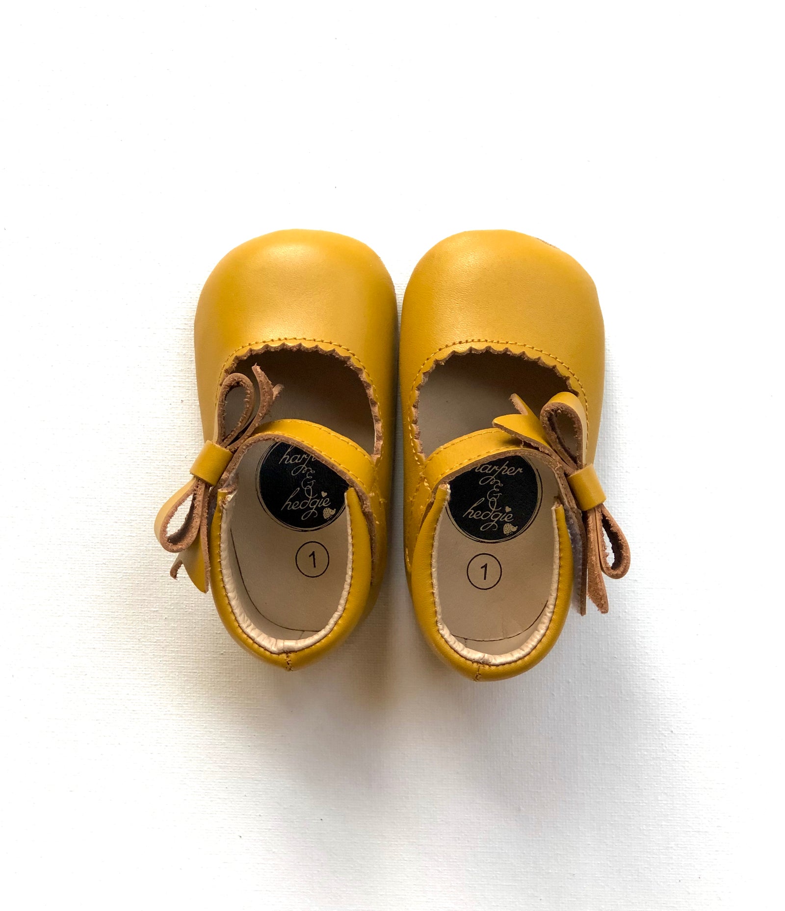 Adorable mustard yellow leather baby Mary Jane shoes with soft soles, light brown suede bows, and “hope & honey” size 1 insole label, displayed on white background.